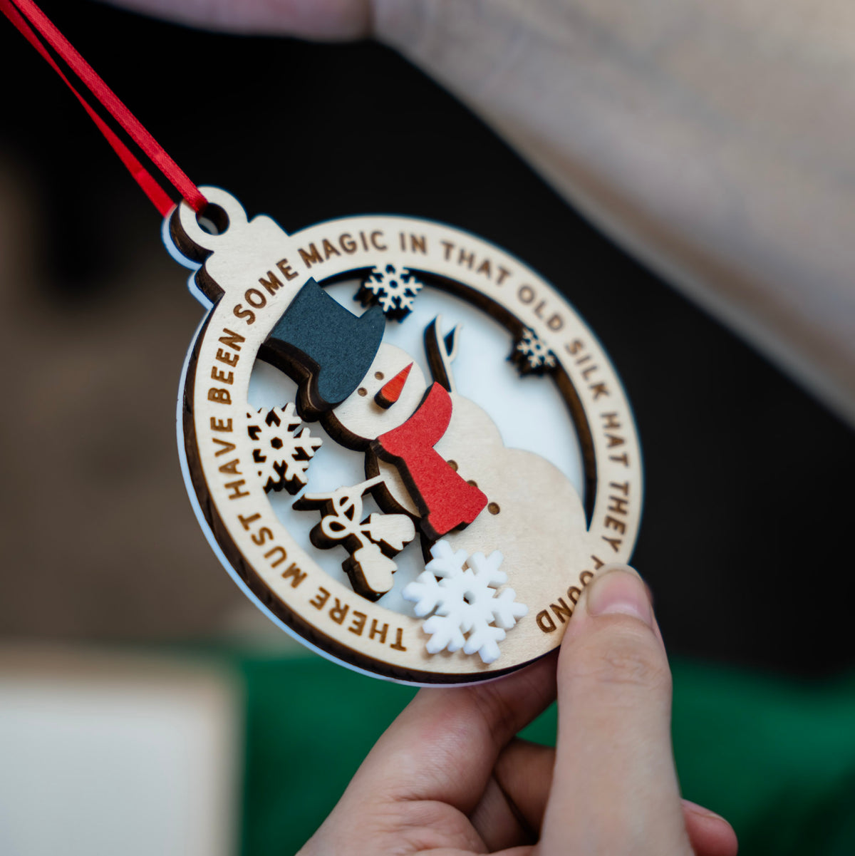 A Close up with a hand that holds a round wooden Christmas ornament featuring a laser-cut and engraved snowman with a red scarf, black hat and carrot nose, surrounded by snowflakes and a red hanging ribbon. The ornament has engraved text around the edge that reads THERE MUST HAVE BEEN SOME MAGIC IN THAT OLD SILK HAT THEY FOUND and is shown against a softly blurred background with green fabric and neutral tones.