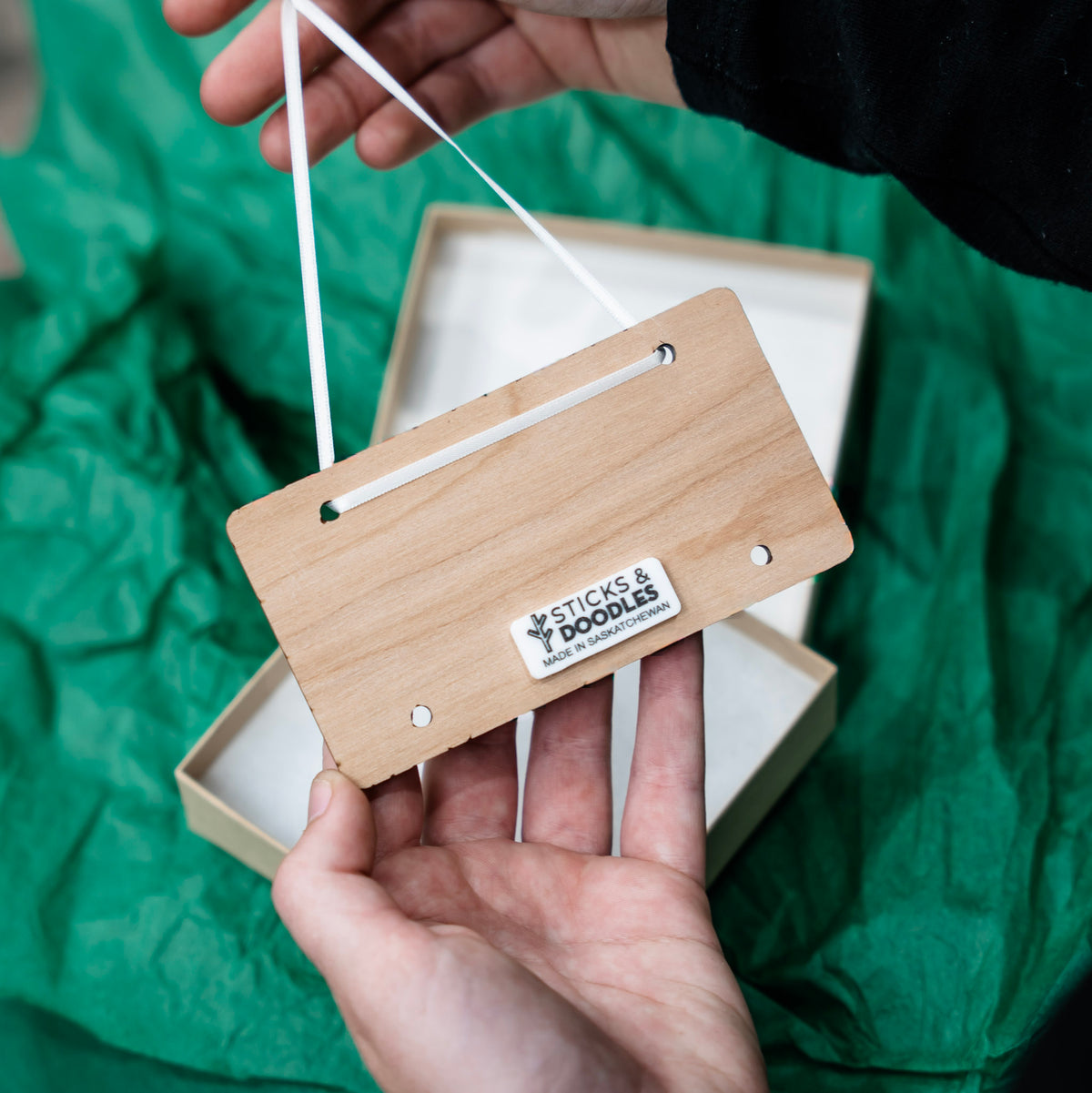 A wooden hanging ornament with a white ribbon is held by hands over a green crinkled fabric background, with an open box visible behind it. The ornament has a visible sticker reading 'STICKS & DOODLES made in saskatchewan'.