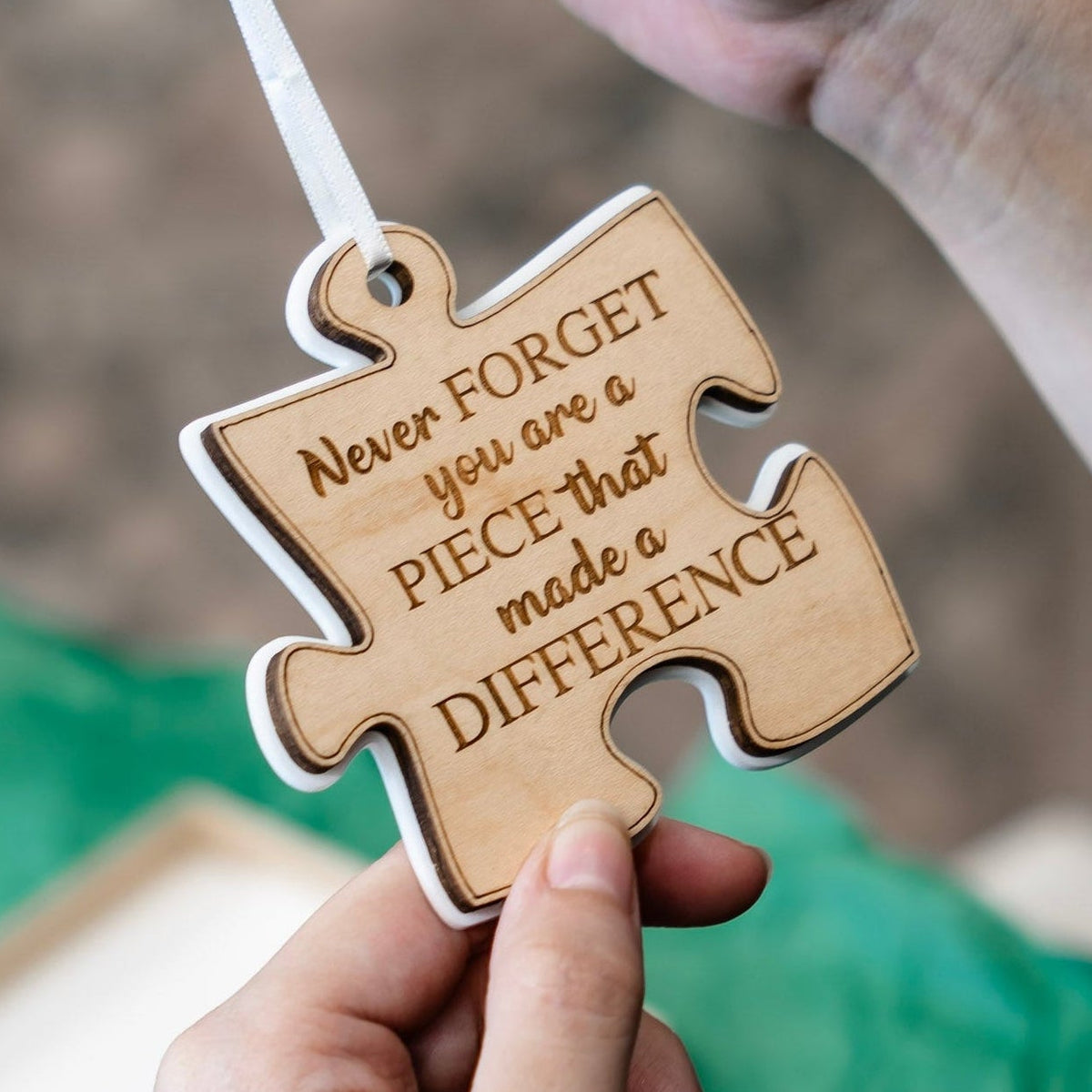 A person holds a wooden puzzle-shaped ornament engraved with the quote 'Never FORGET you are a PIECE that made a DIFFERENCE' on a white ribbon, with white acrylic backer with a blurred green gift bag in the background.
