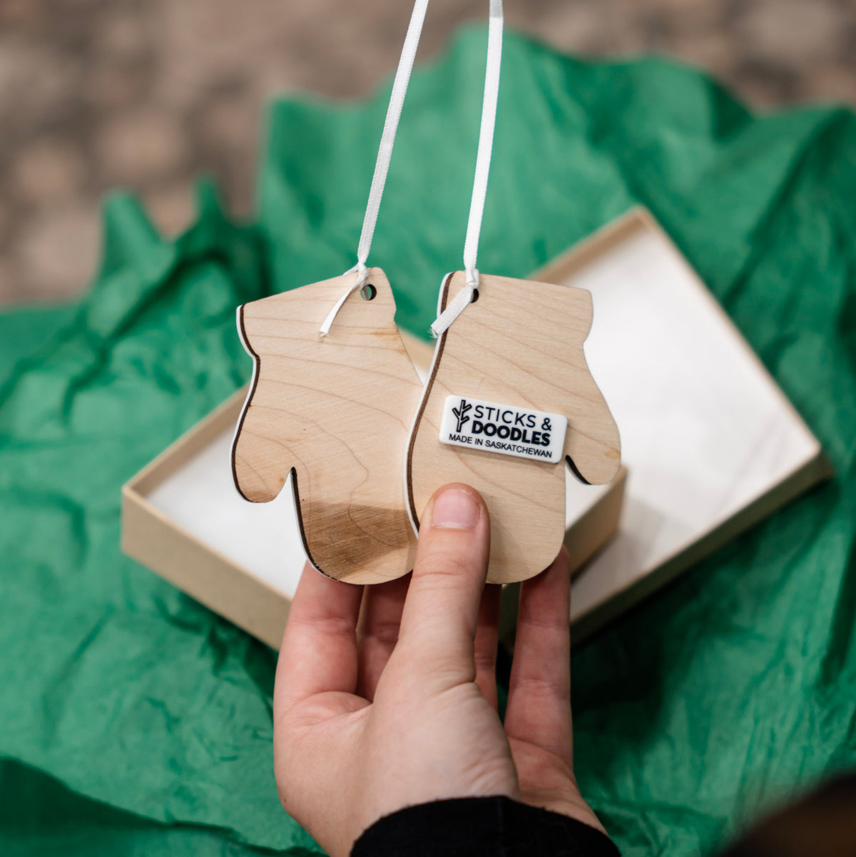 A hand holds two small wooden ornament shapes with white hanging ribbon. One ornament has a visible label reading 'STICKS & DOODLES MADE IN Saskatchewan'.