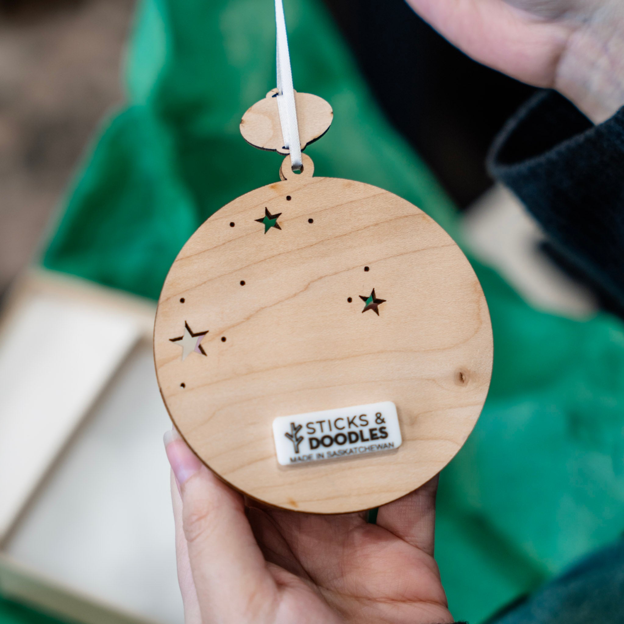 A person holds a round wooden hanging ornament with star and dot cutouts and a visible 'STICKS & DOODLES' label. The background shows a green fabric surface and part of a hand holding the item.