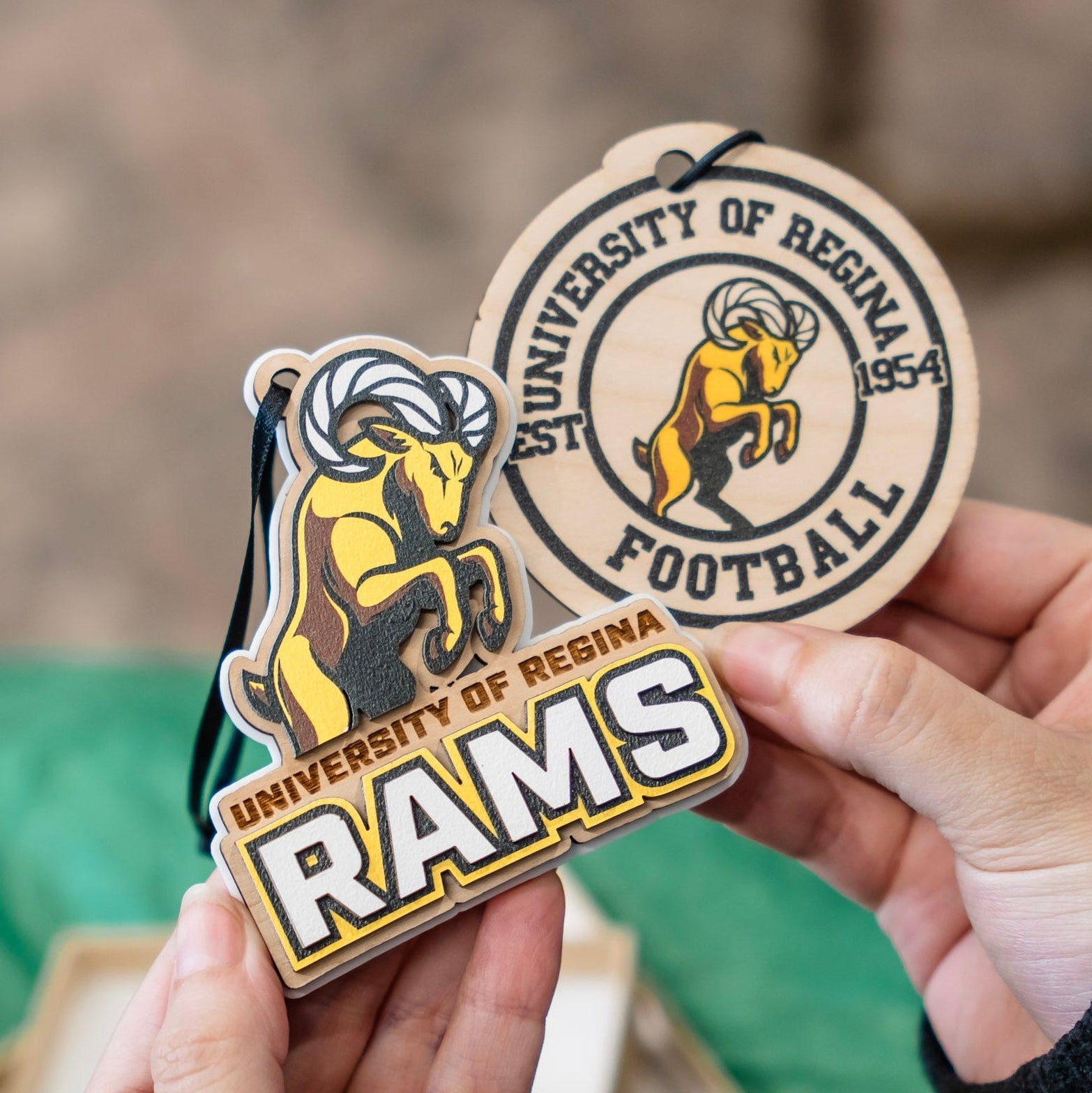 Two University of Regina Rams ornament and car charm held in hands: a round wooden football tag and a matching wooden ram-shaped ornament, both featuring the yellow ram logo and school name.