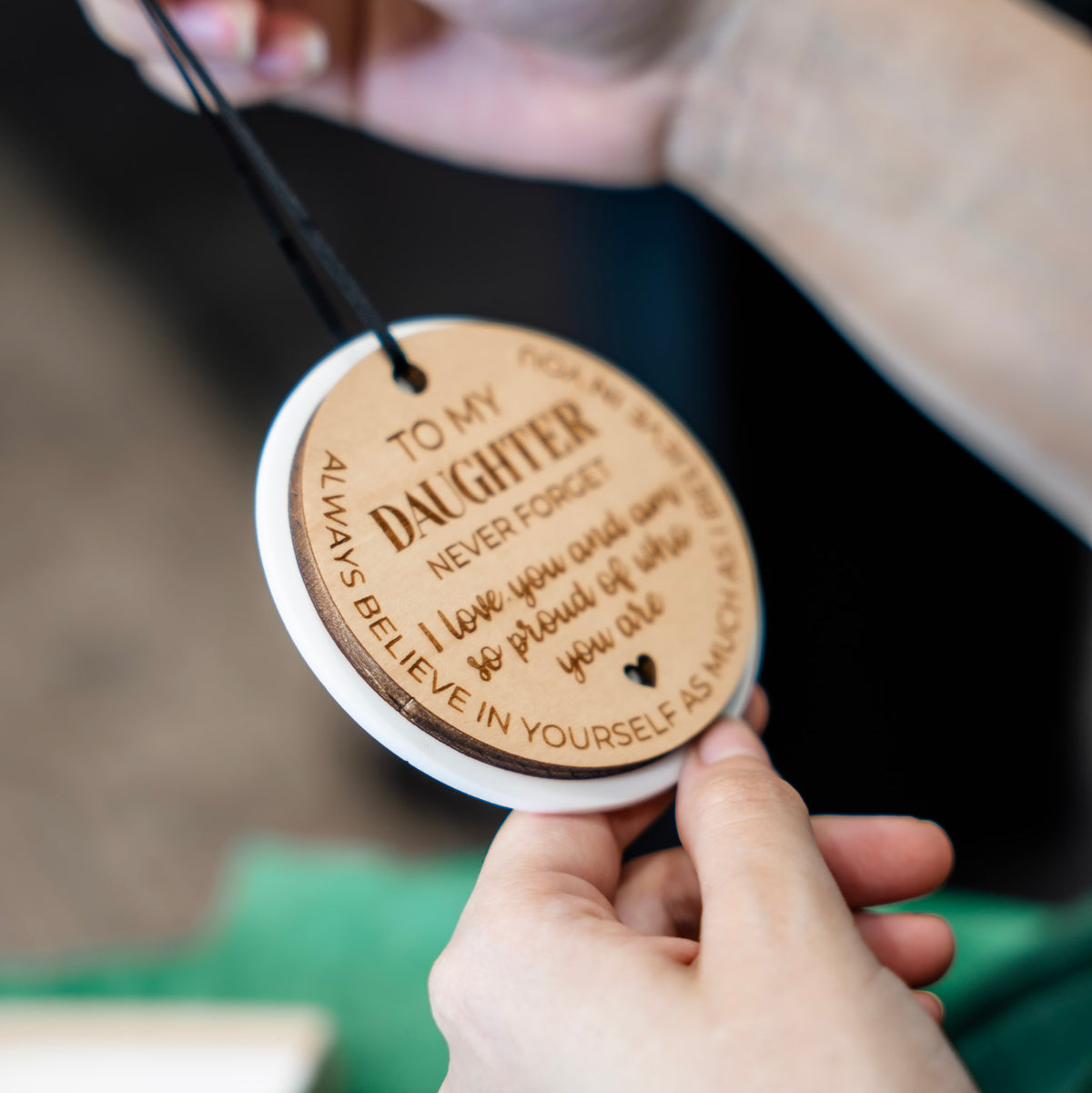 A side view of a round wooden ornament with engraved text for a daughter. The ornament is light brown wood with engraved dark brown lettering and a black ribbon, shown against a blurred background with green tissue paper.