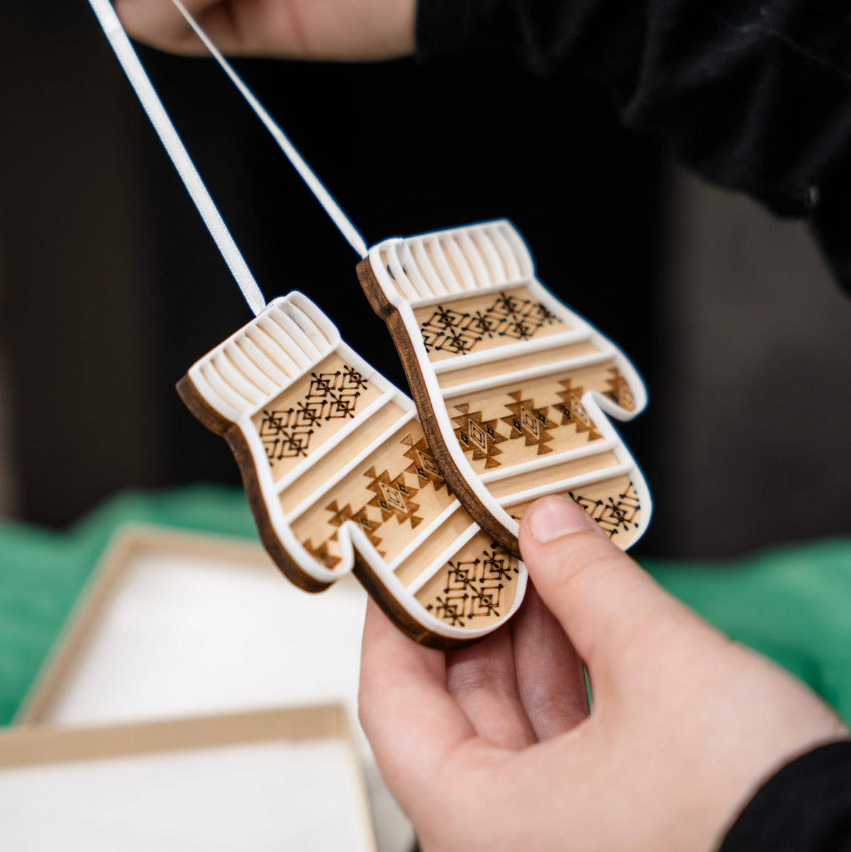 A hand holds two wooden Christmas ornament shaped like mittens with a laser-engraved and scored geometric pattern. The ornaments are suspended by white ribbon, with a gift box and bright green tissue paper in the background.
