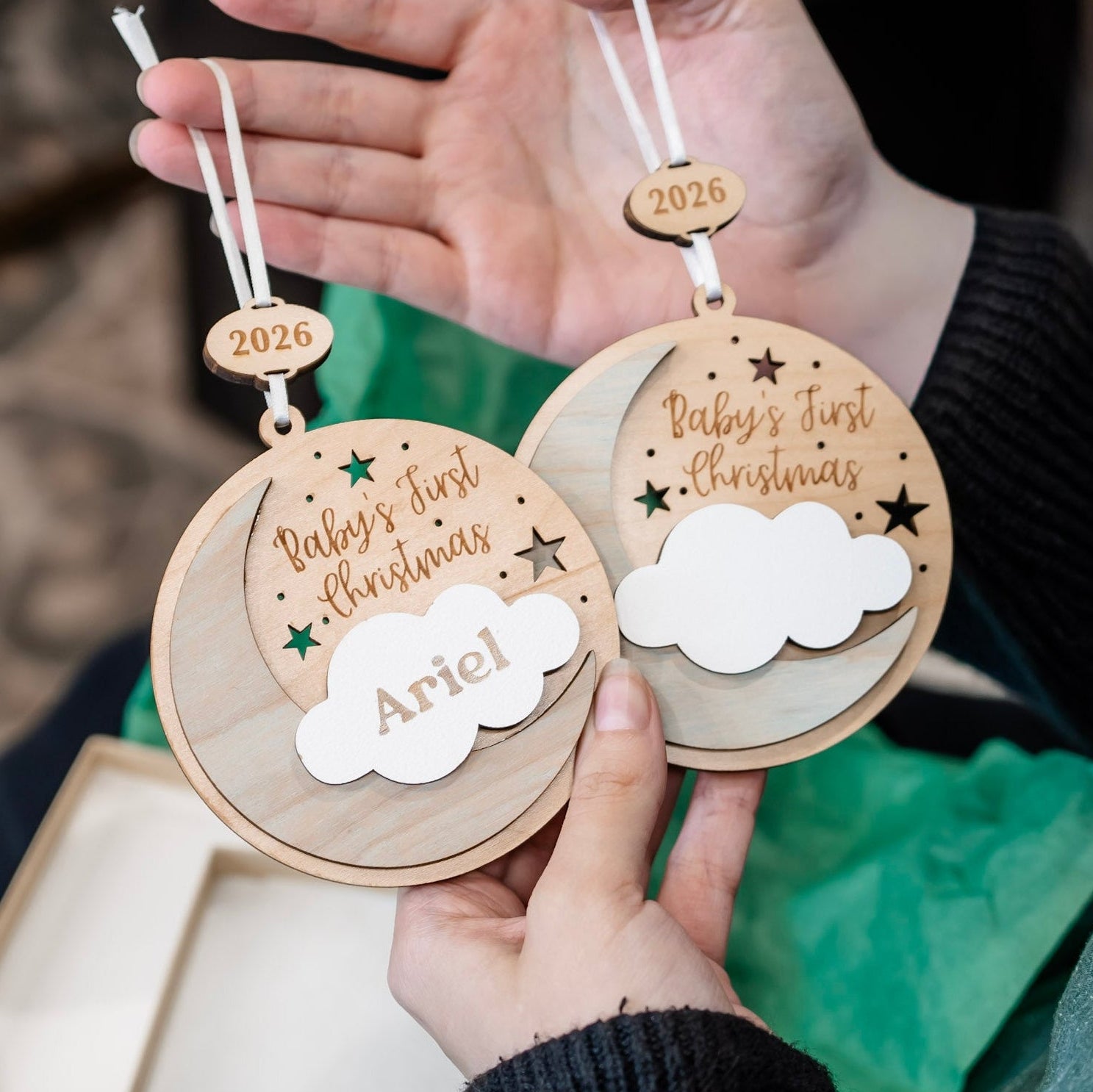 Two personalized wooden Christmas tree ornaments held in hands, featuring laser-engraved crescent moons, stars, and the text 'Baby's First Christmas' with the name 'Ariel' on a white cloud cutout. Each ornament has a small wooden tag engraved with '2026' and a white hanging string.