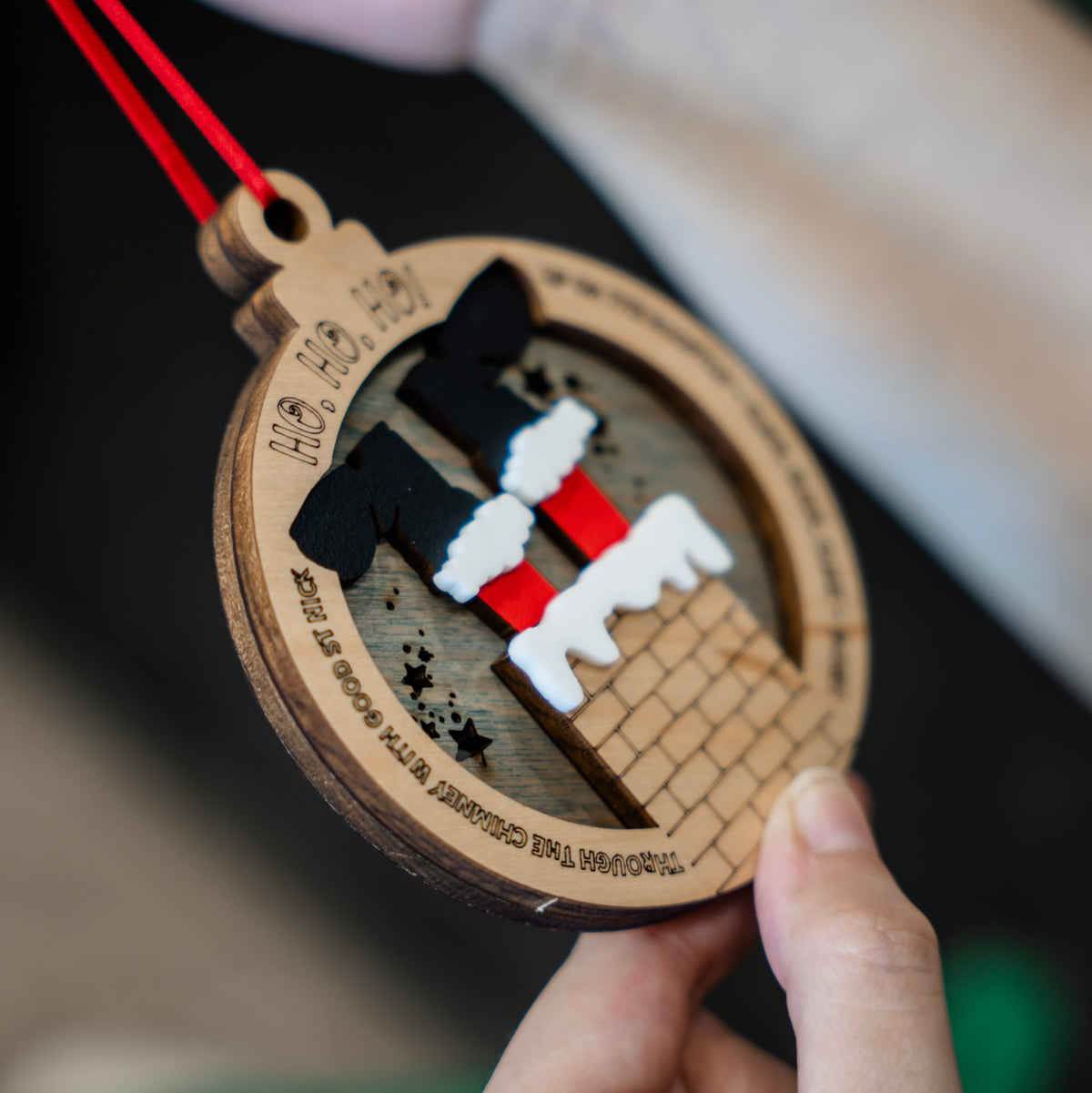 A close-up of a hand holding a round wooden Christmas ornament while another hand holds the red hanging ribbon. The ornament features a chimney with two legs in black and red socks with white trim, set against a stained blue wood background with the text 'HO HO HO!' and the song Up on the rooftop Christmas song.