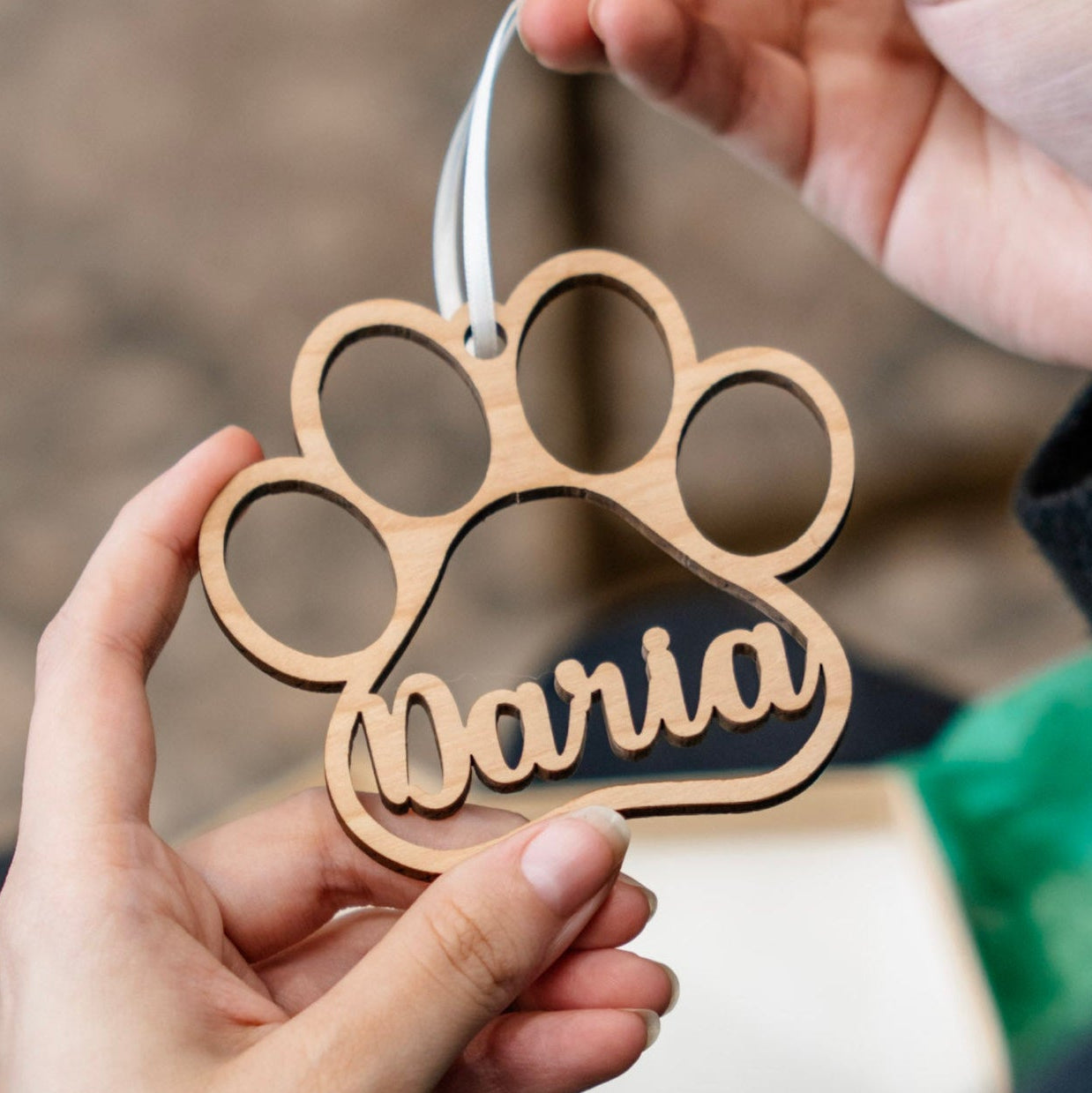 A wooden paw print hanging ornament with the name 'Daria' cut out in the center, held by two hands against a softly blurred indoor background.