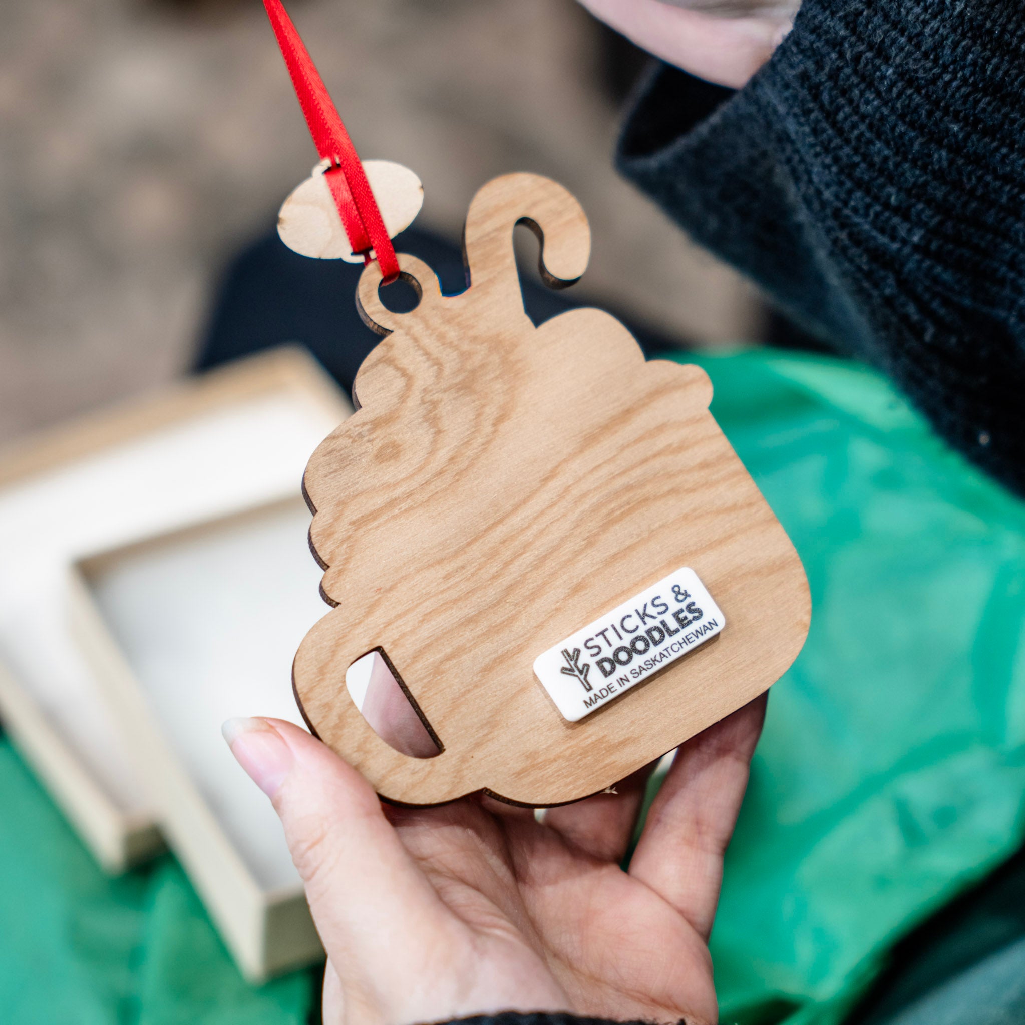 A hand holds a wooden mug-shaped ornament with a red hanging ribbon. The natural wood grain is visible, and a small white sticker on the front reads 'Cicks & Doodles'.