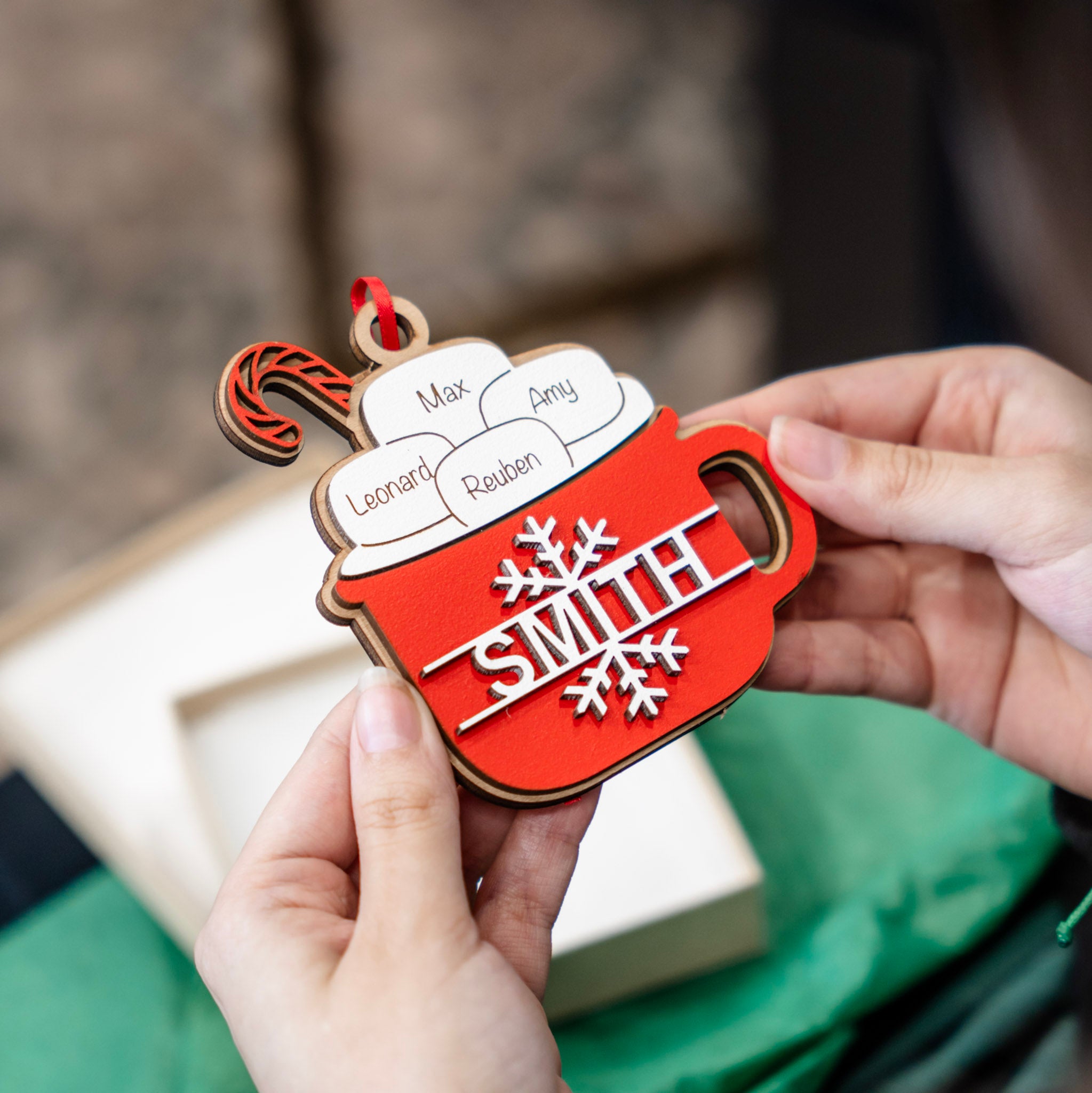 A close-up of hands holding a red and white personalized holiday ornament shaped like a hot cocoa mug, with the name 'SMITH' and snowflake details, against a softly blurred background.