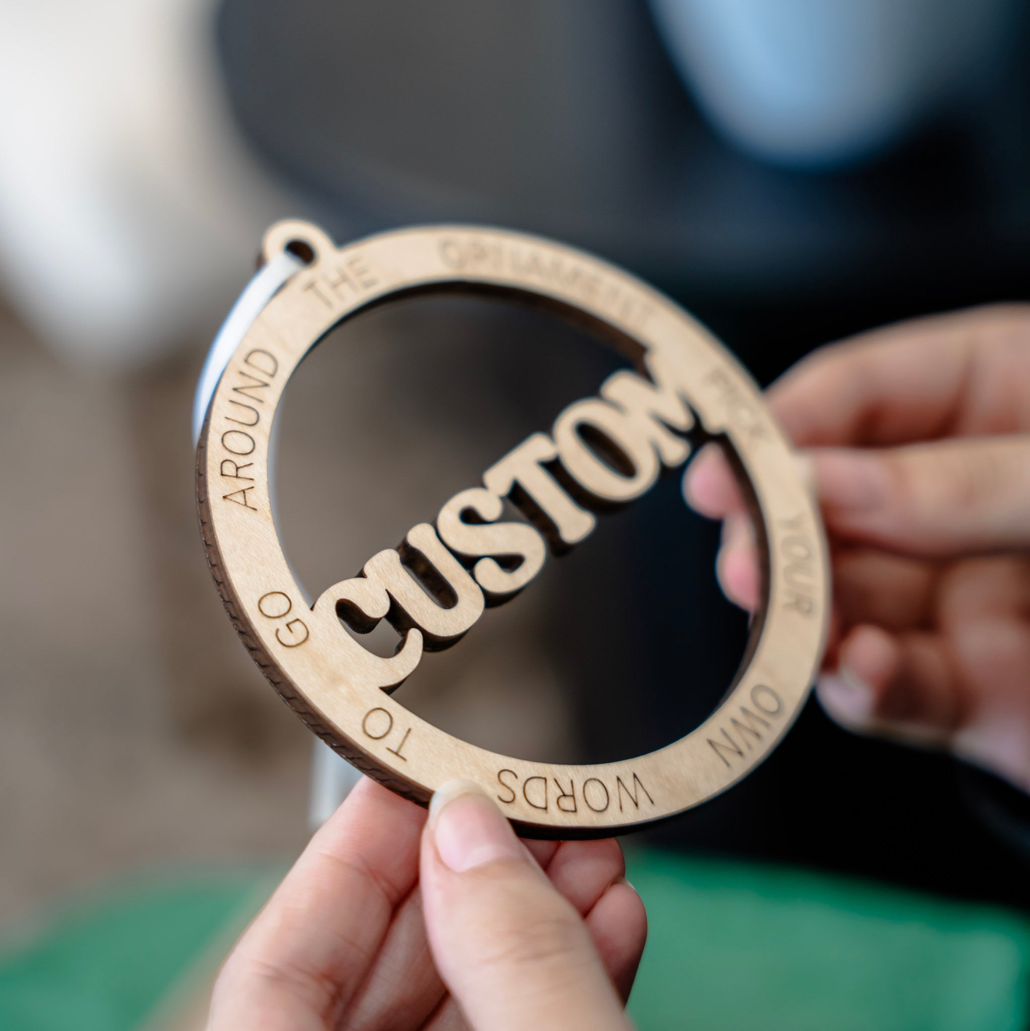A close-up side view of a person holding a circular wooden ornament or ornament with the word 'CUSTOM' in the center and engraved text around the border.