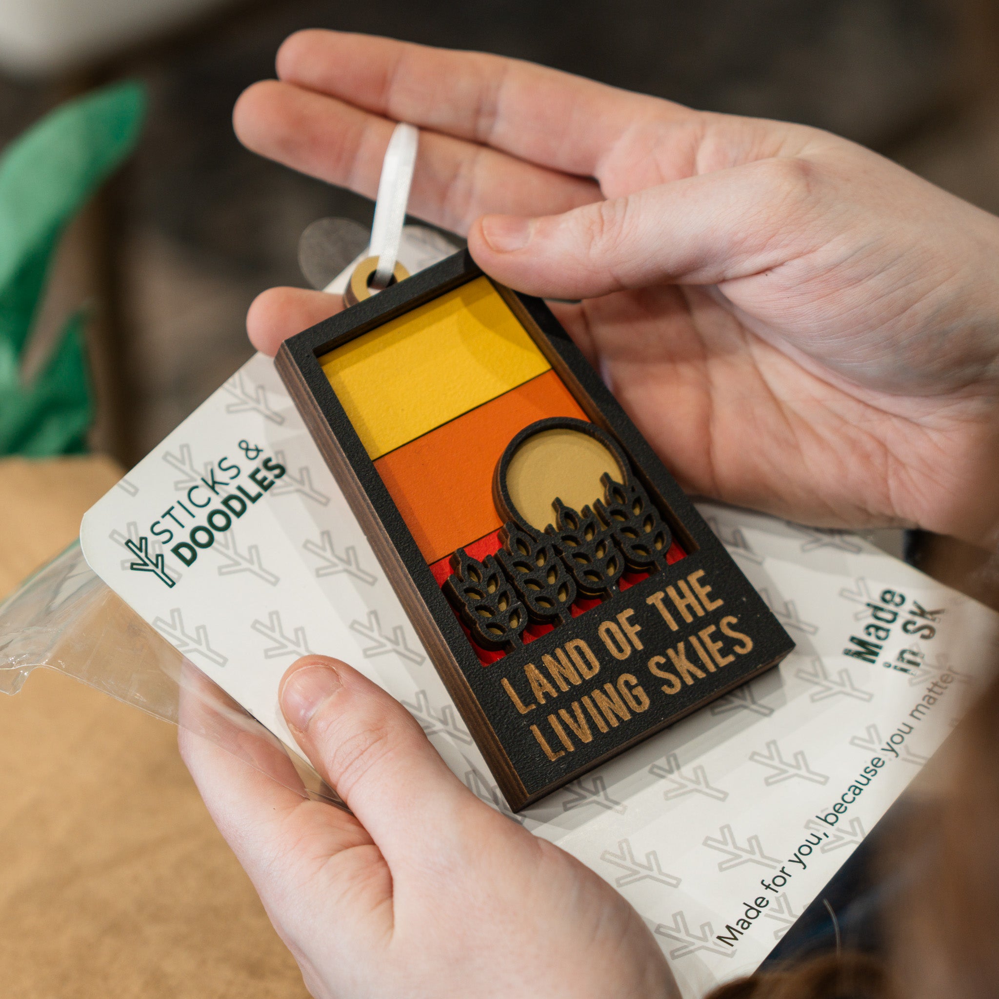 A person holds a rectangular wooden ornament or tag with a white hanging loop. The tag has a black border and reads “LAND OF THE LIVING SKIES,” with a stylized sunset design in yellow, orange, and brown, plus small black tree silhouettes.