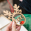 A wooden reindeer-shaped Christmas ornament with a red hanging ribbon, held by hands over a gift box. The ornament features a laser-cut reindeer head silhouette with the name 'Leo' in cursive across the face and a small red wooden nose.