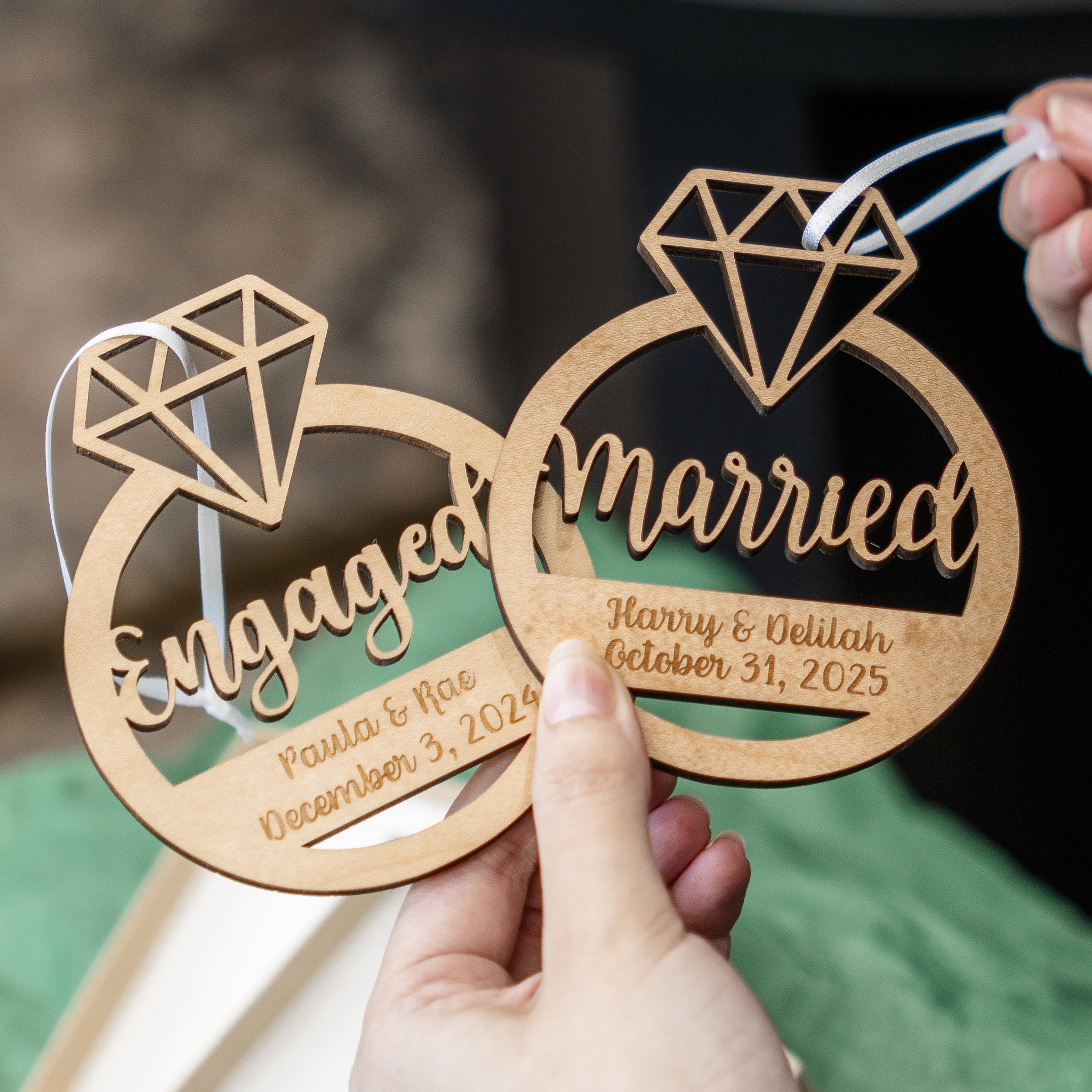 Two personalized wooden ring-shaped ornaments being held in hands, one engraved with 'Engaged' and the other with 'married', both featuring a diamond outline at the top and custom names and dates.