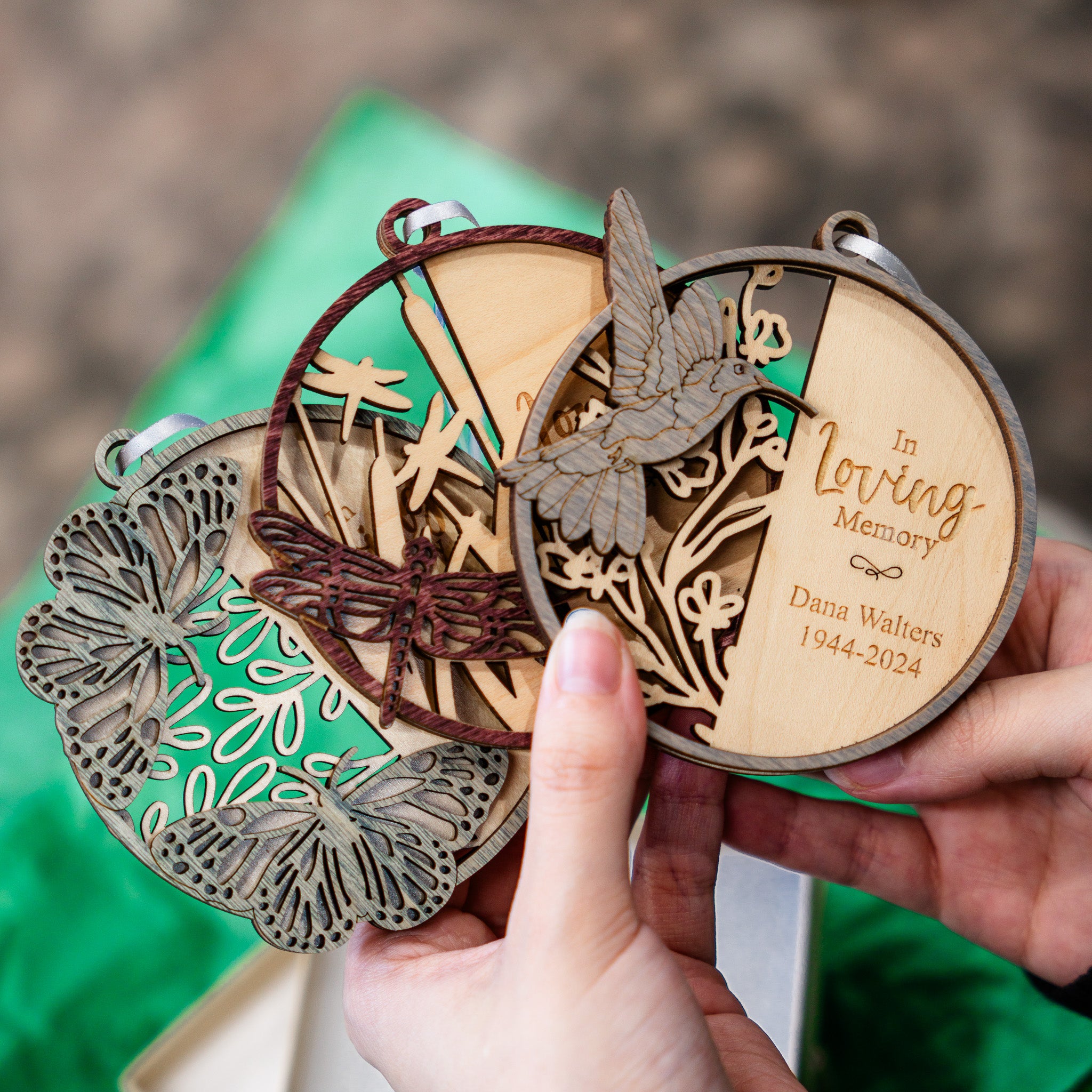 A person holds three personalized laser-cut wooden keepsake ornaments with butterfly, dragonfly, and bird designs over a green gift box. One ornament is engraved with the text 'In Loving Memory Dana Walters 1944-2024'.