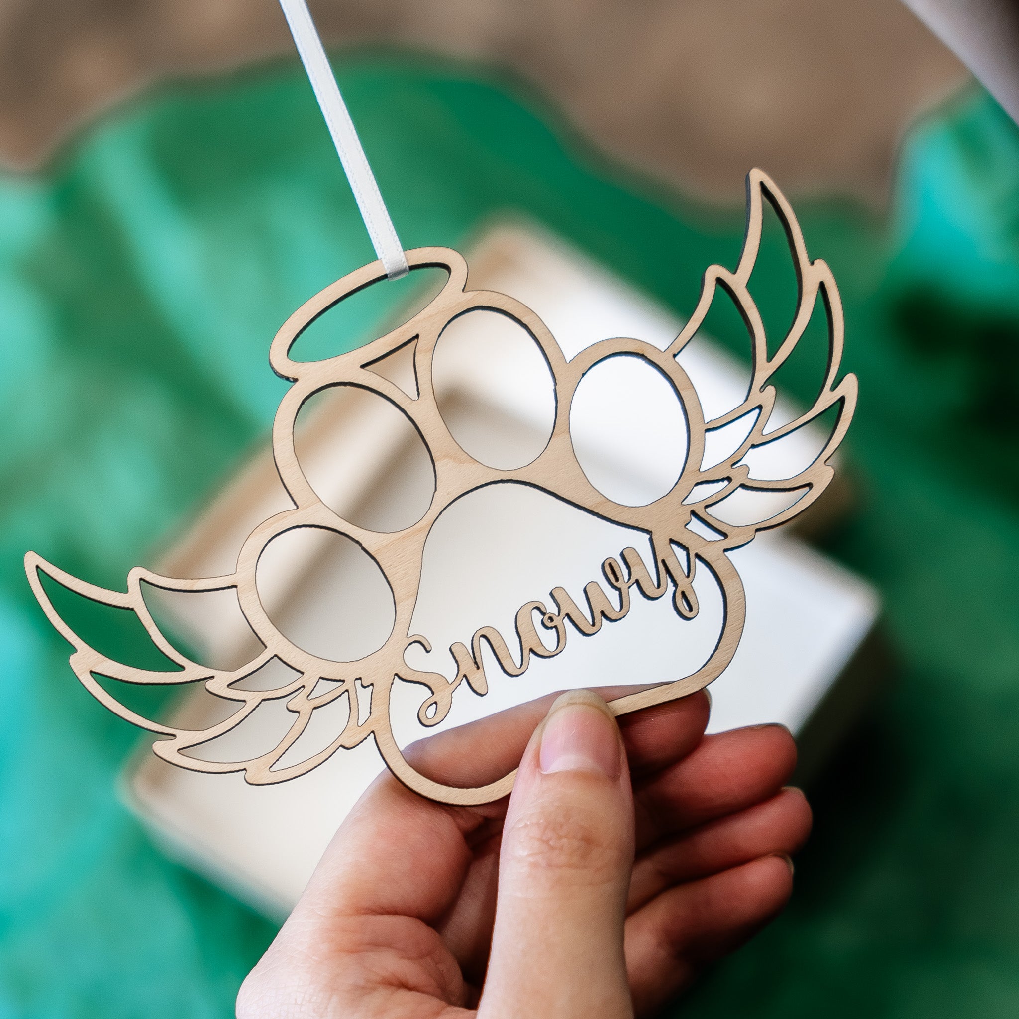 A hand holds a laser-cut wooden paw print ornament with wings and the name 'Snowy' cut into the center, set against a blurred green and white background.