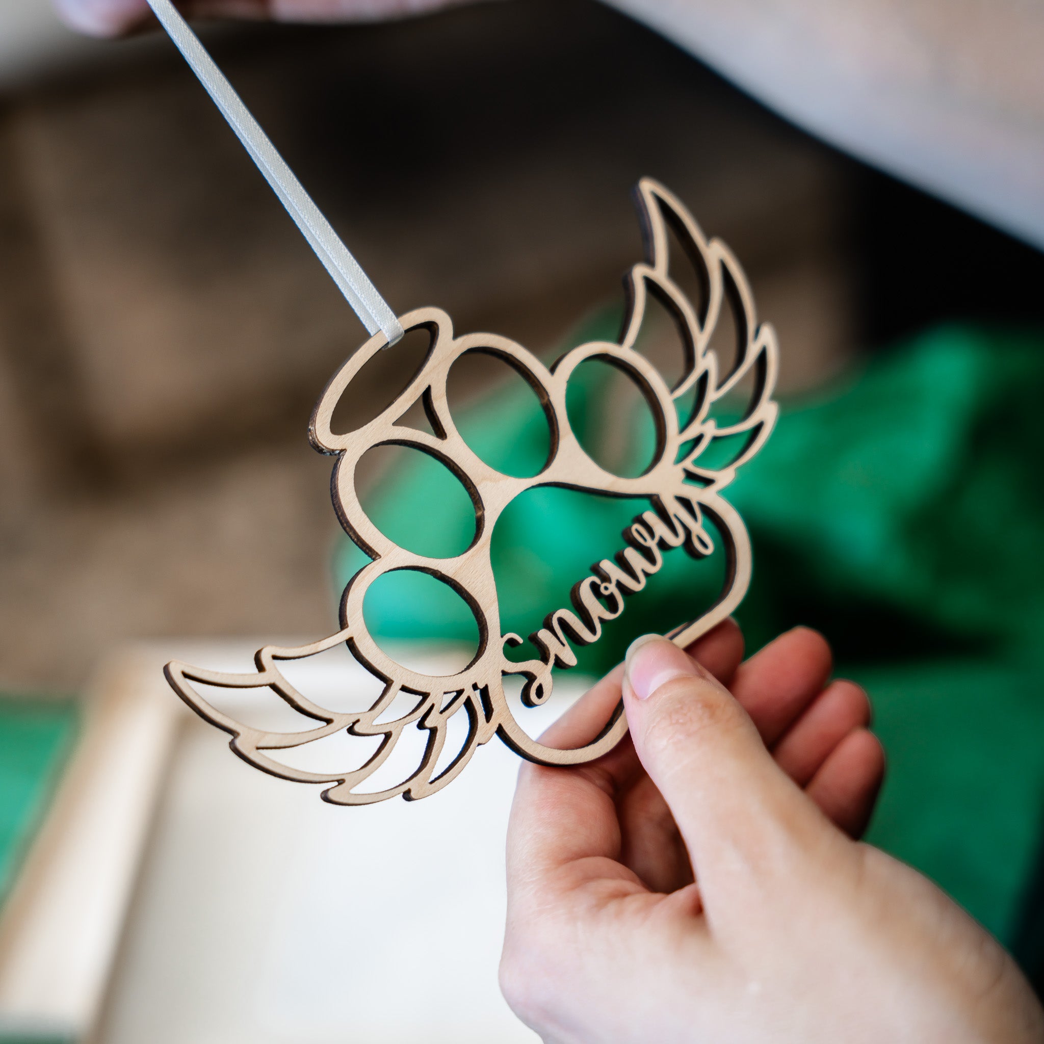 A hand holds a laser-cut wooden paw print decoration with wings and the word 'snowy' in script, against a softly blurred background with green fabric.