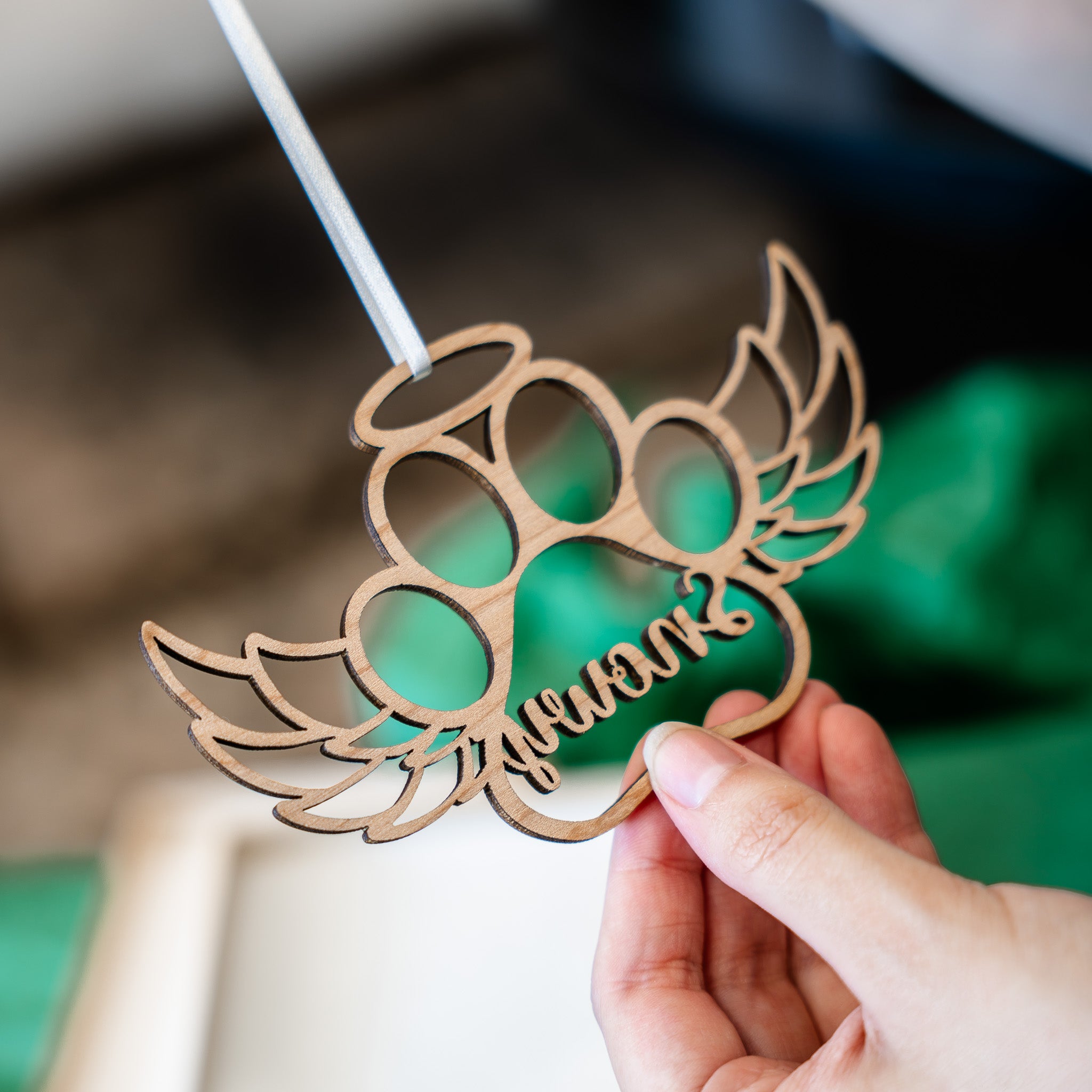 A hand holding a laser-cut wooden angel wings decoration with the name 'Quinn' cut out in the center, set against a blurred background.