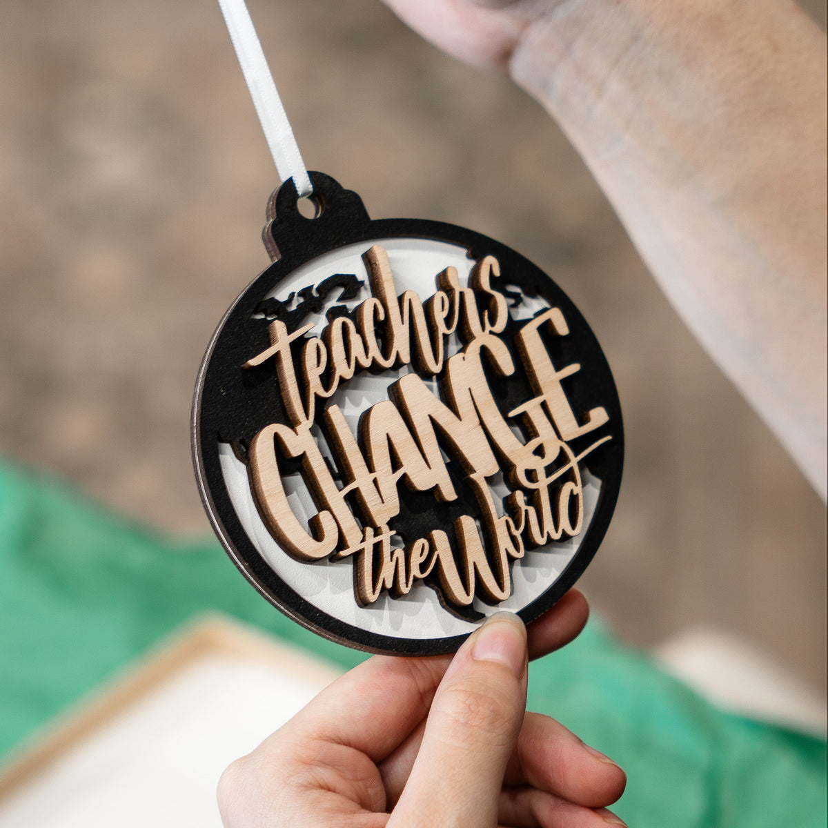 A round wooden ornament held by a hand, featuring the laser-cut text 'teacher's CHANCE the World' in light wood over a black and white background.