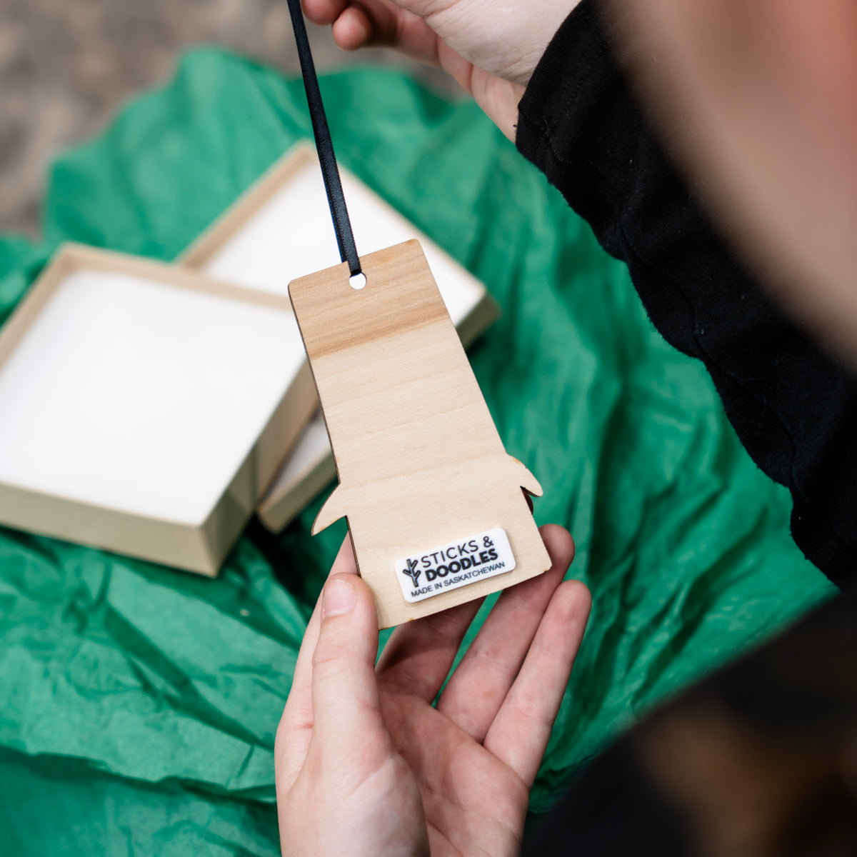 A person holds a light natural wood ornament tag with a black ribbon. The tag has a small white sticker at the bottom with the brand name 'STICKS & DOODLES MADE IN SASKATCHEWAN'.