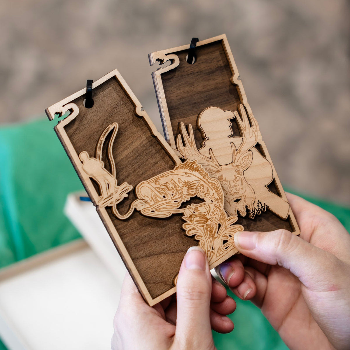 A close-up of hands holding two rectangular wooden ornaments with laser-cut fish and deer designs, with green tissue paper and an gift box in the background.