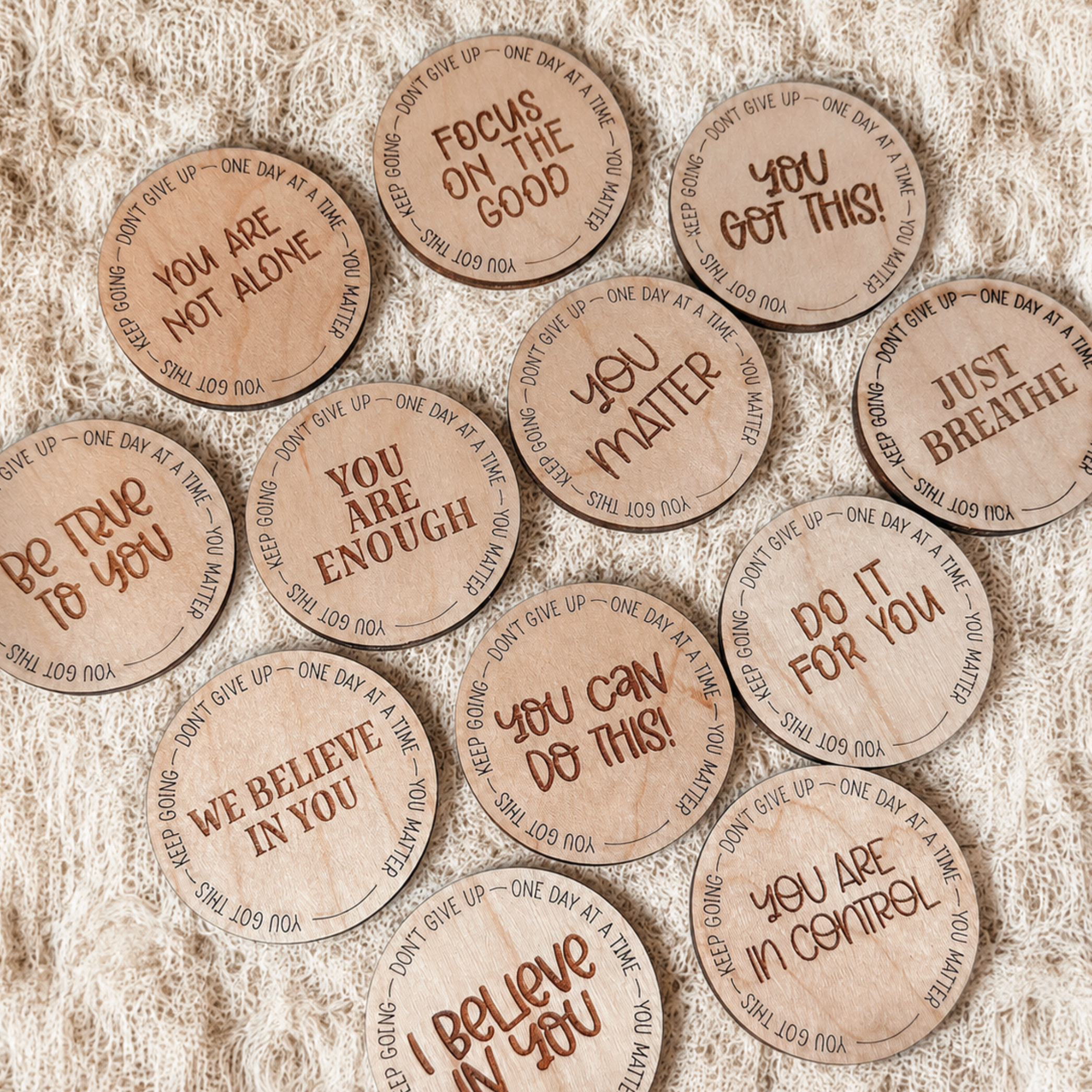 A group of round wooden tokens or coasters with engraved positive affirmations, arranged on a textured beige fabric background.