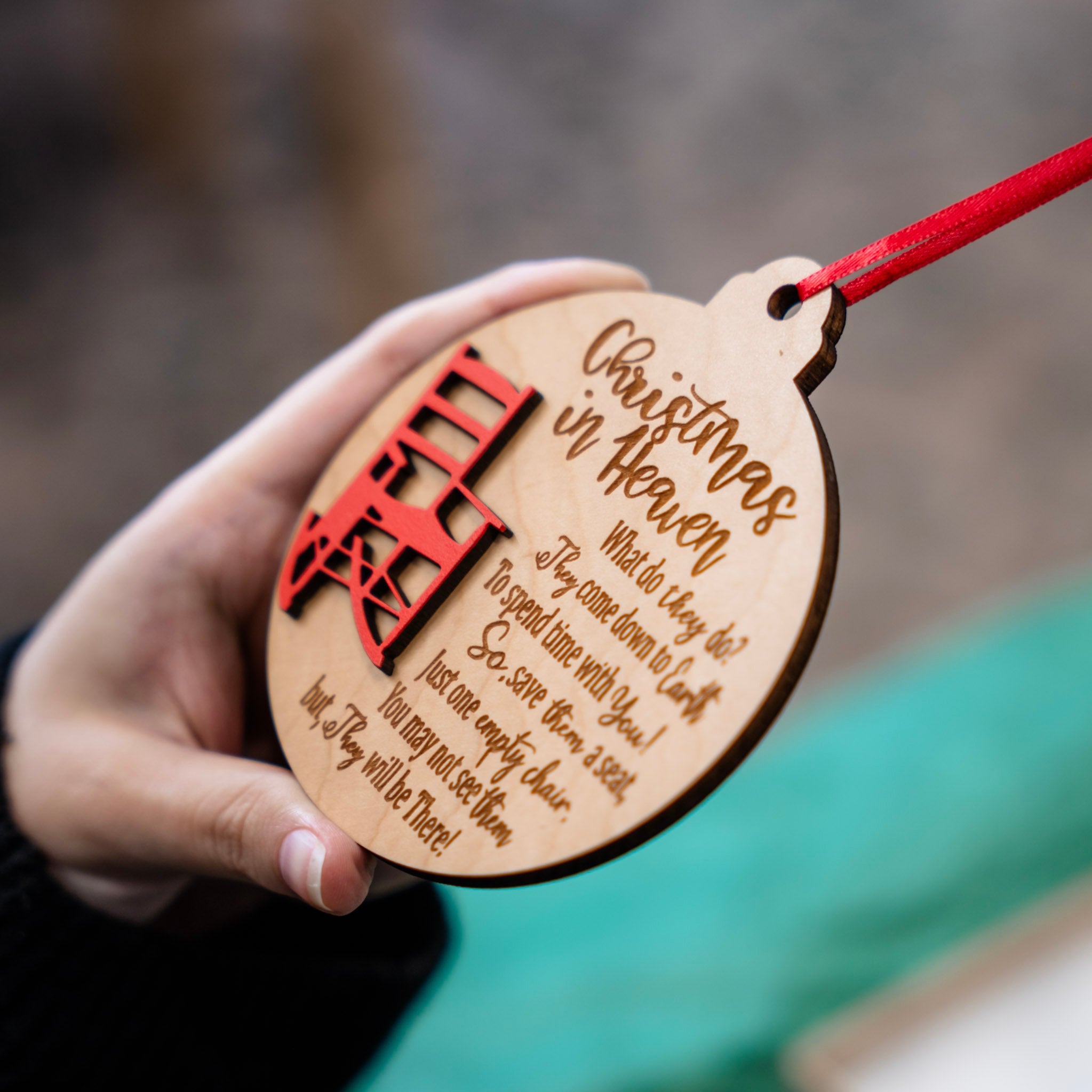 A hand holds a round wooden Christmas ornament engraved with 'Christmas in Heaven' and a red hanging ribbon, with a blurred outdoor background.