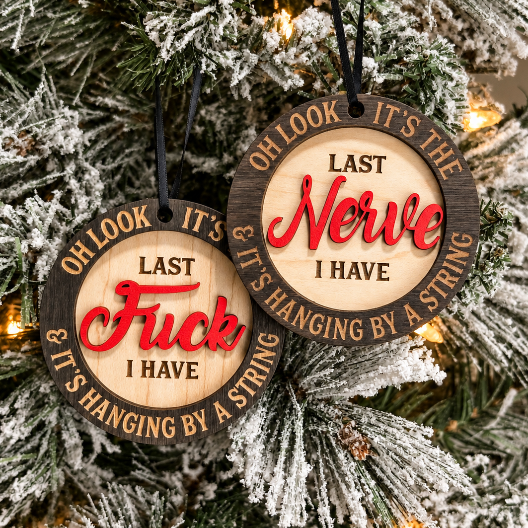 Two round wooden Christmas ornaments hanging on a frosted Christmas tree. The ornaments have a natural wood grain border, engraved black text, and red script text reading 'OH LOOK IT'S THE LAST NERVE I HAVE' and 'OH LOOK IT'S THE LAST Fuck I HAVE & IT'S HANGING BY A STRING'.