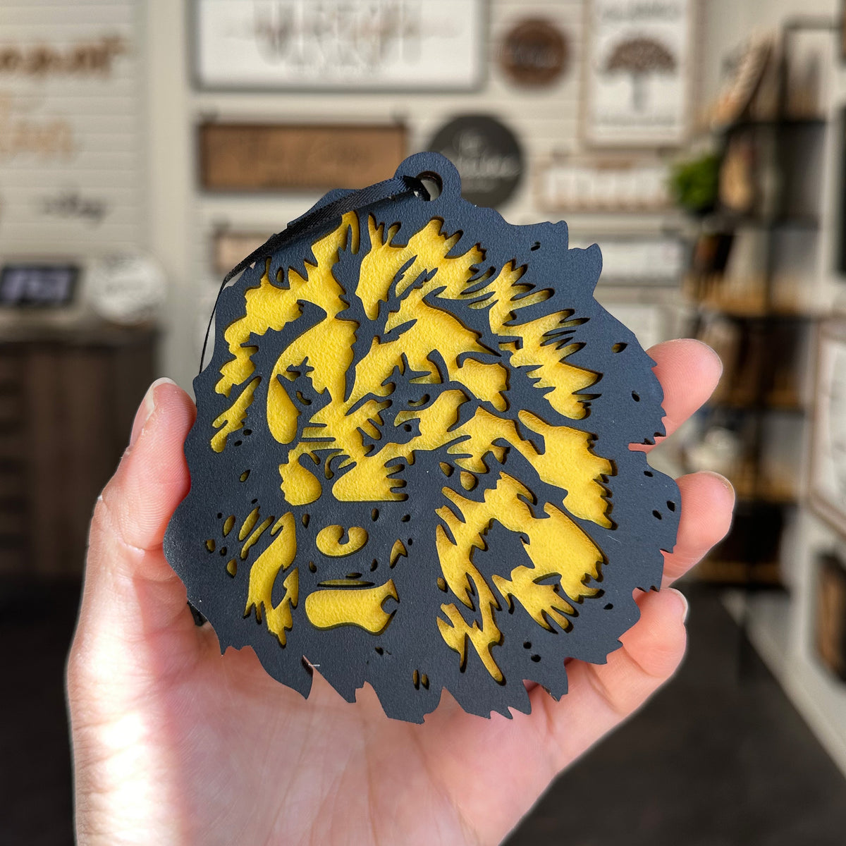 A hand holds a round black and yellow laser-cut wood ornament featuring a stylized lion face, with a blurred shop interior in the background.