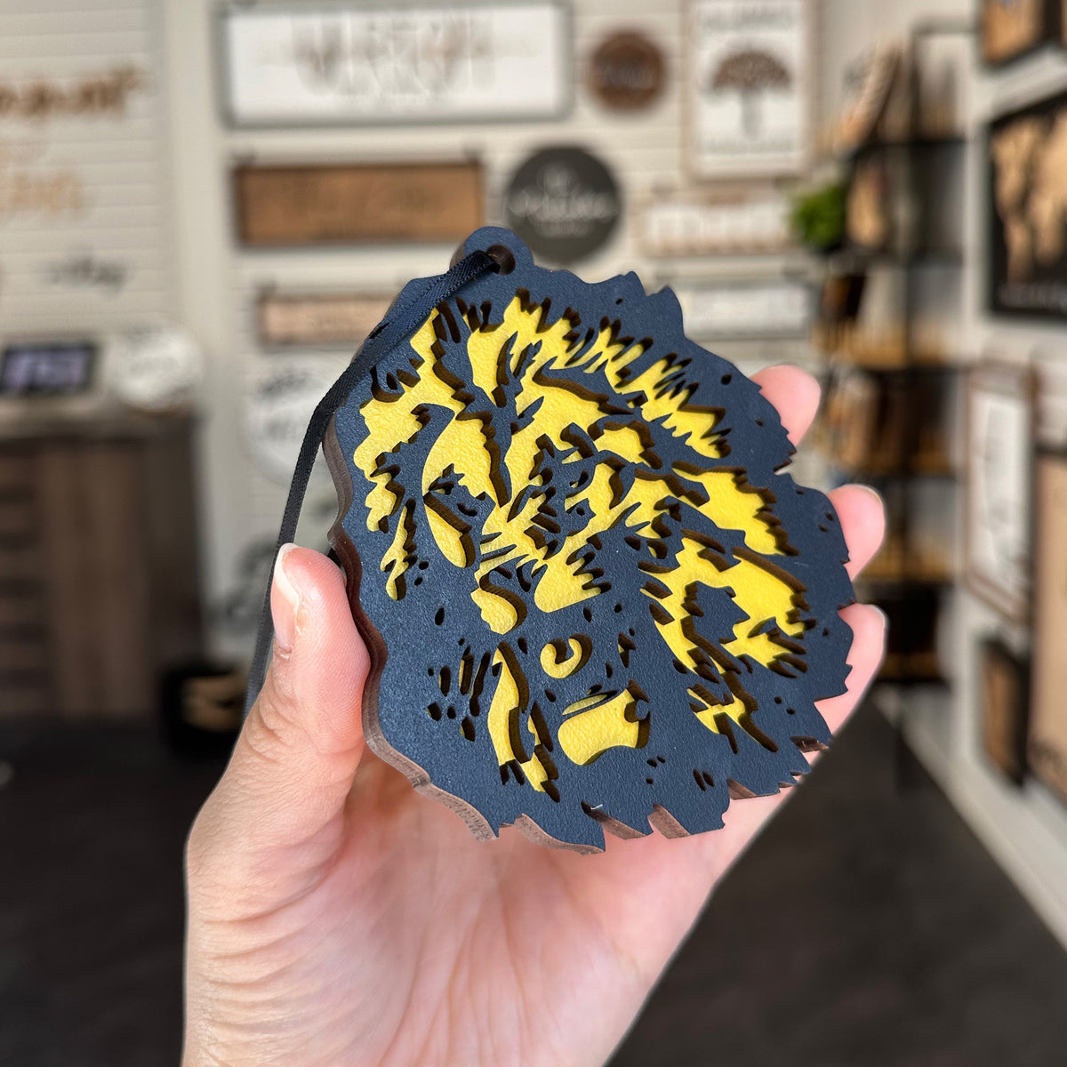 A hand holding a round black and yellow laser-cut wooden ornament featuring a stylized tree design, with a blurred shop interior in the background.