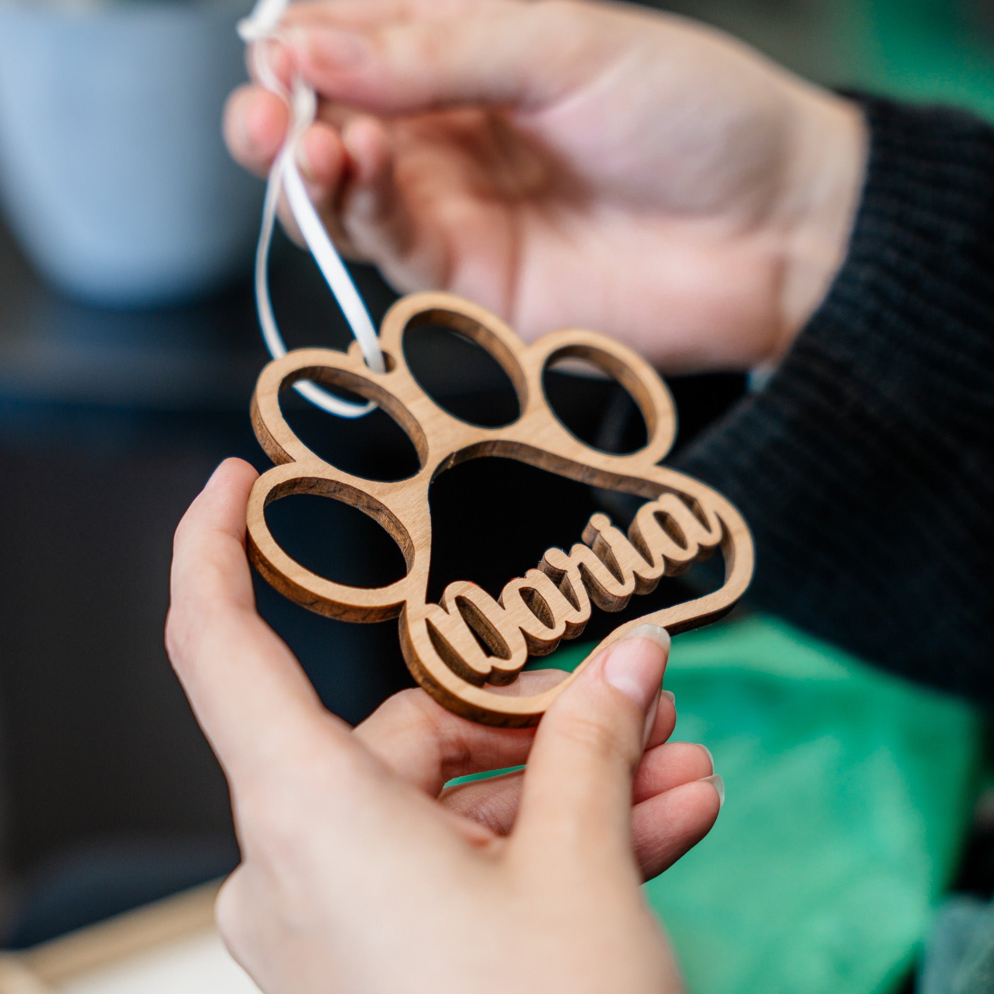 A wooden paw-shaped hanging ornament with the name 'Nadia' cut into the bottom, held by two hands. The ornament is made of light brown wood and hangs from a white string.